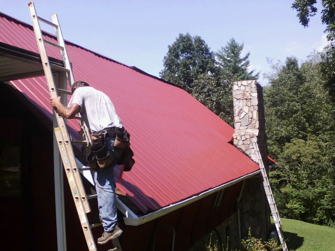 Roofer on a ladder beside a red metal roof and stone chimney — Soffit & Fascia Repair by Roofing Brothers in Elmwood